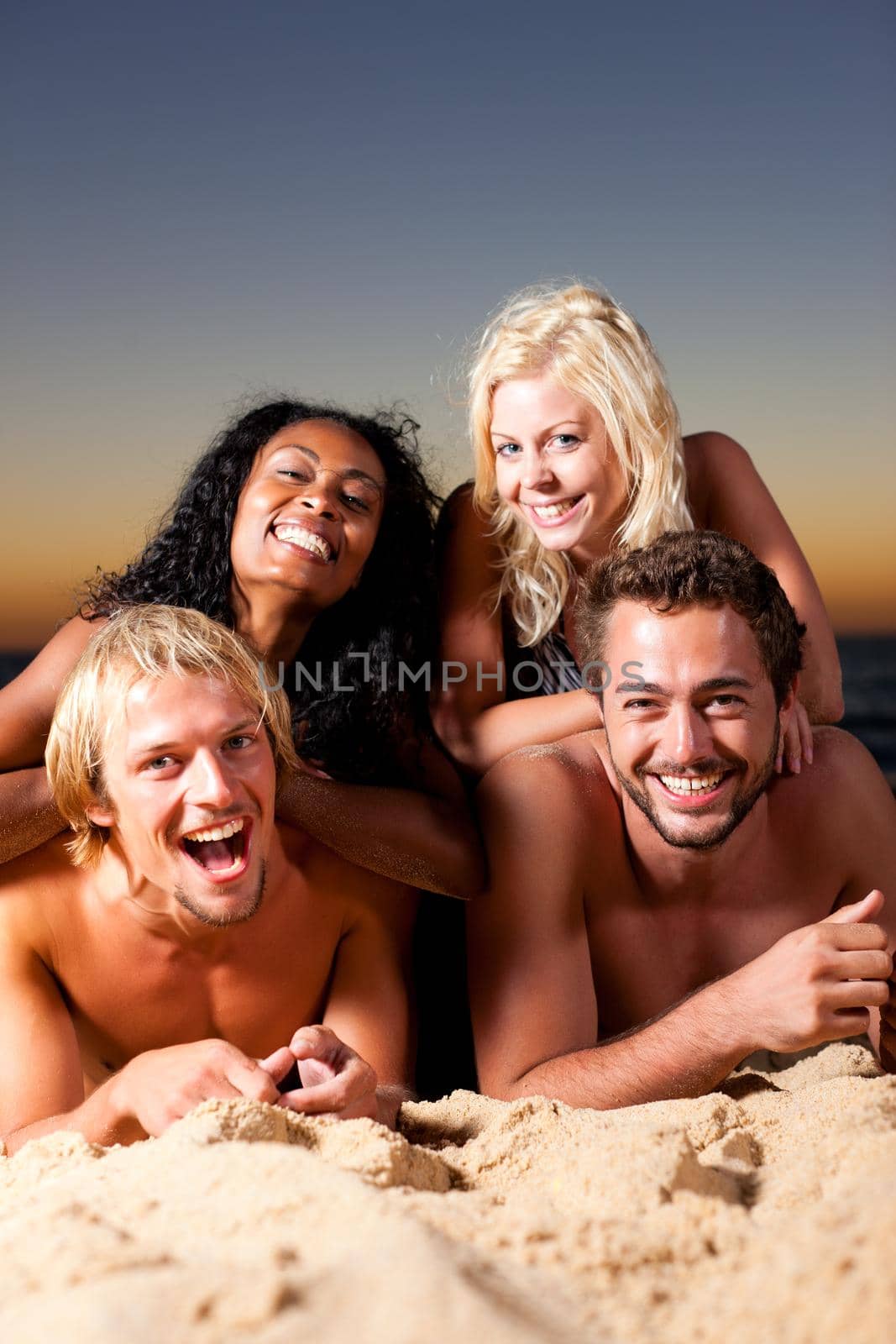 Group of four friends - men and women - sitting on the beach against the afterglow of a sunset over the ocean