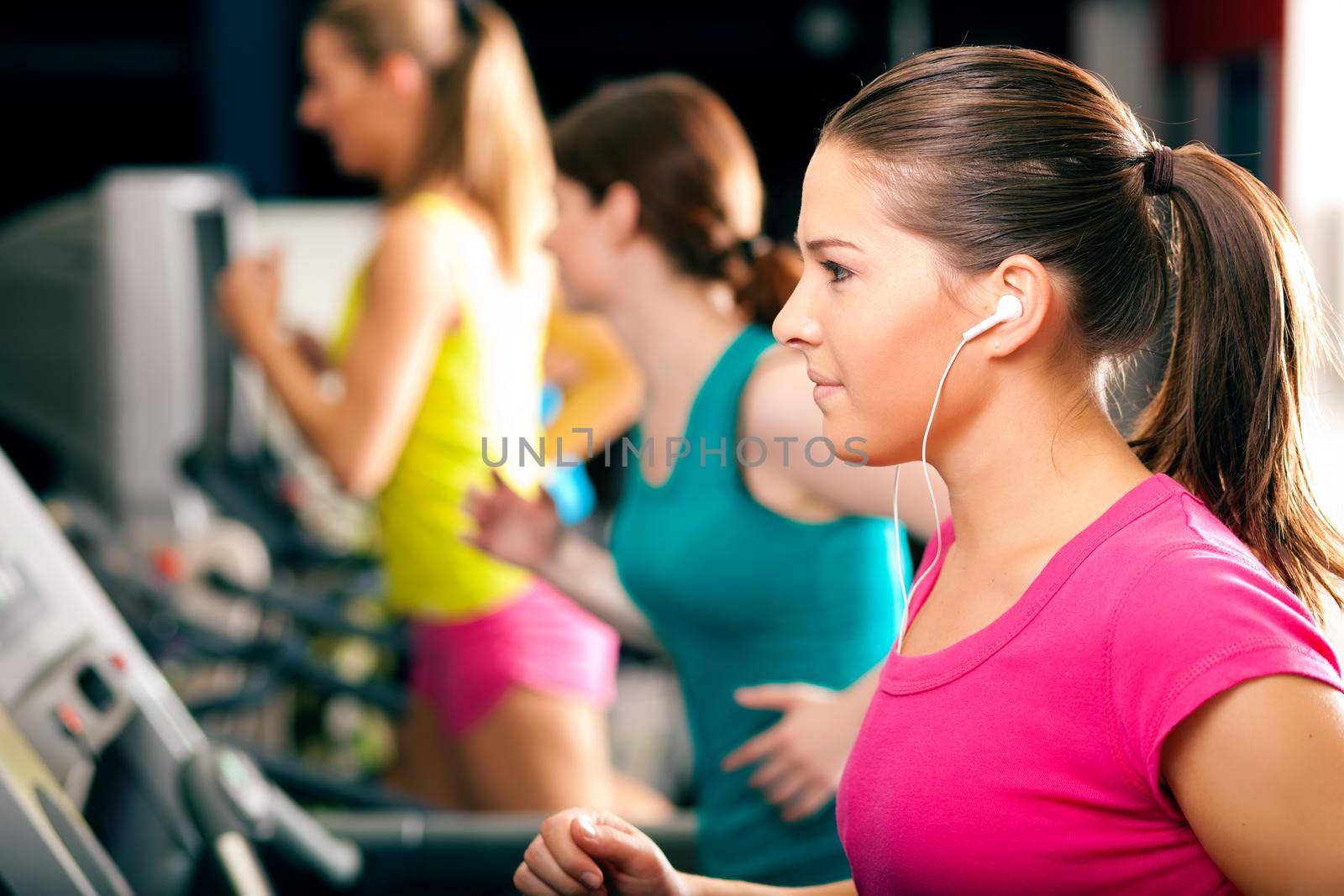 Running on treadmill in gym - group of women exercising to gain more fitness, the woman in front wears earplugs and enjoys music