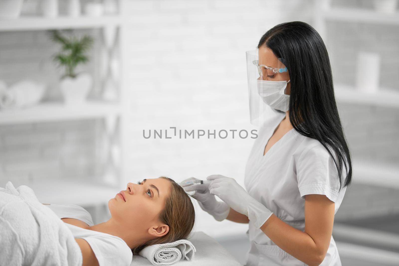 Side view of brunette young beautician sitting and doing special injection for growth hair in beauty room. Concept of procedure for hair restoration and improvements.