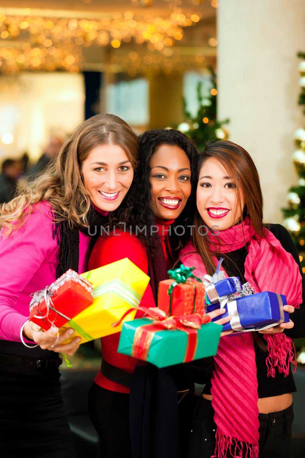 Group of three women - white, black and Asian - with Christmas presents in a shopping mall in front of a Christmas tree