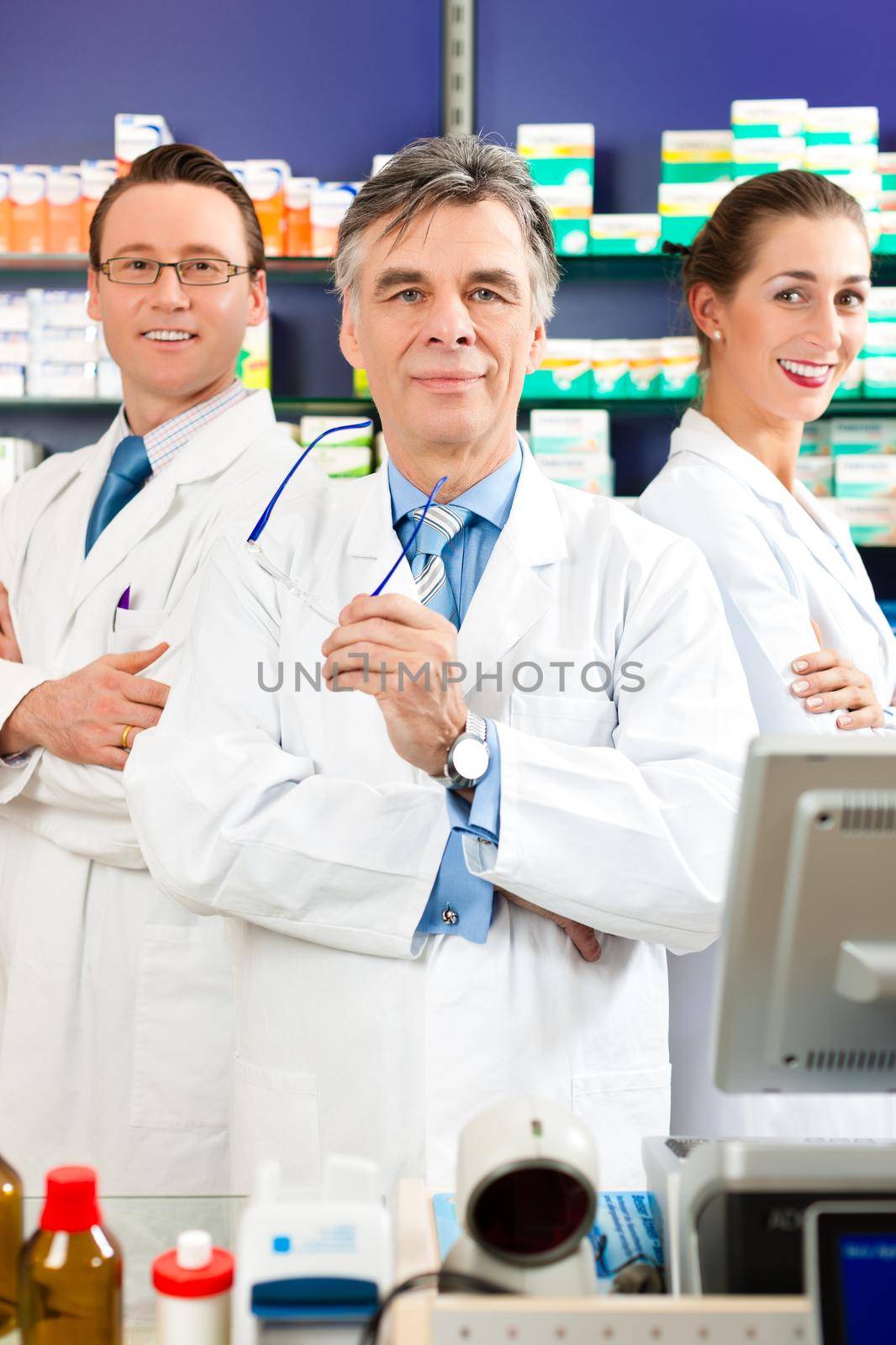 Pharmacist with his team standing in pharmacy or drugstore in front of shelves with pharmaceuticals