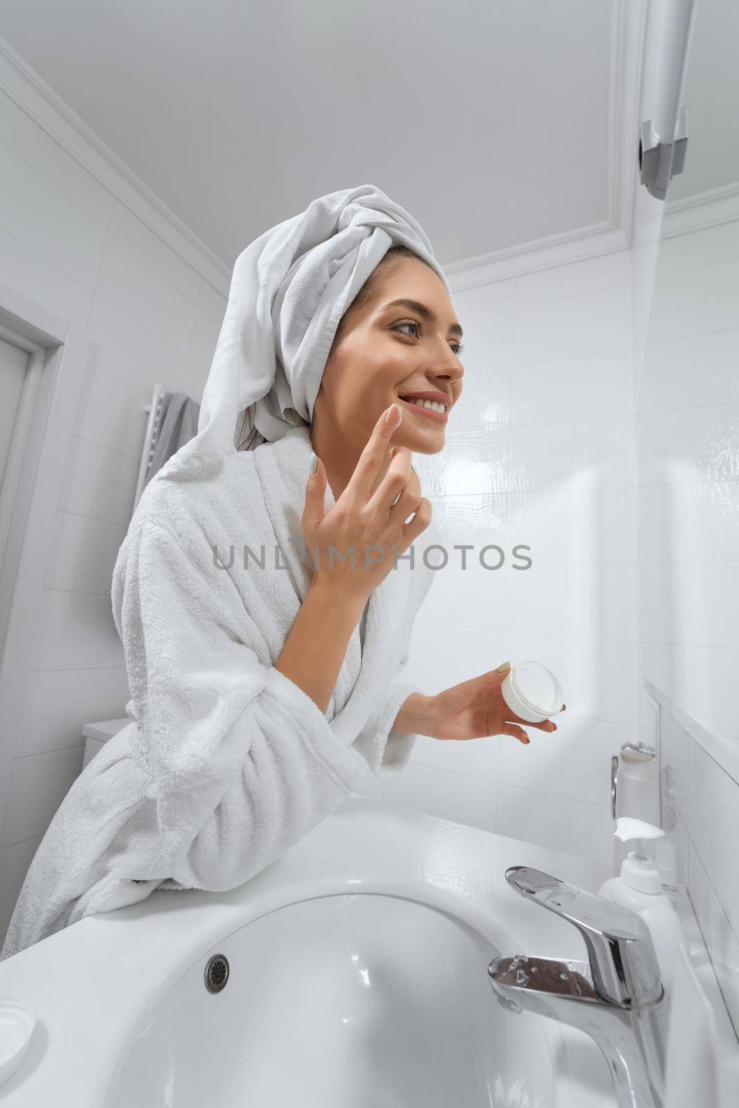 Side view of smiling beautiful young woman looking at mirror and using face cream in white robe. Concept of time for yourself and care body with good mood at home.