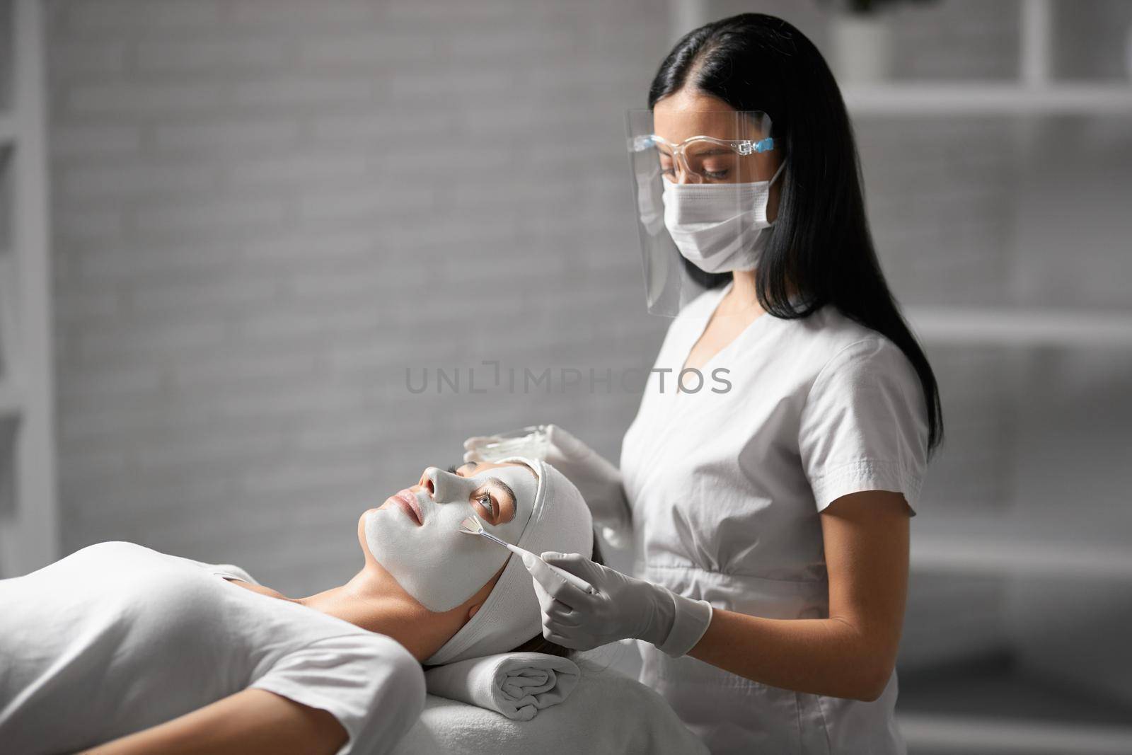 Side view of beautiful brunette beautician in protective mask doing peeling face for young attractive woman. Concept of procedure for improvements skin with special professional cosmetology. 