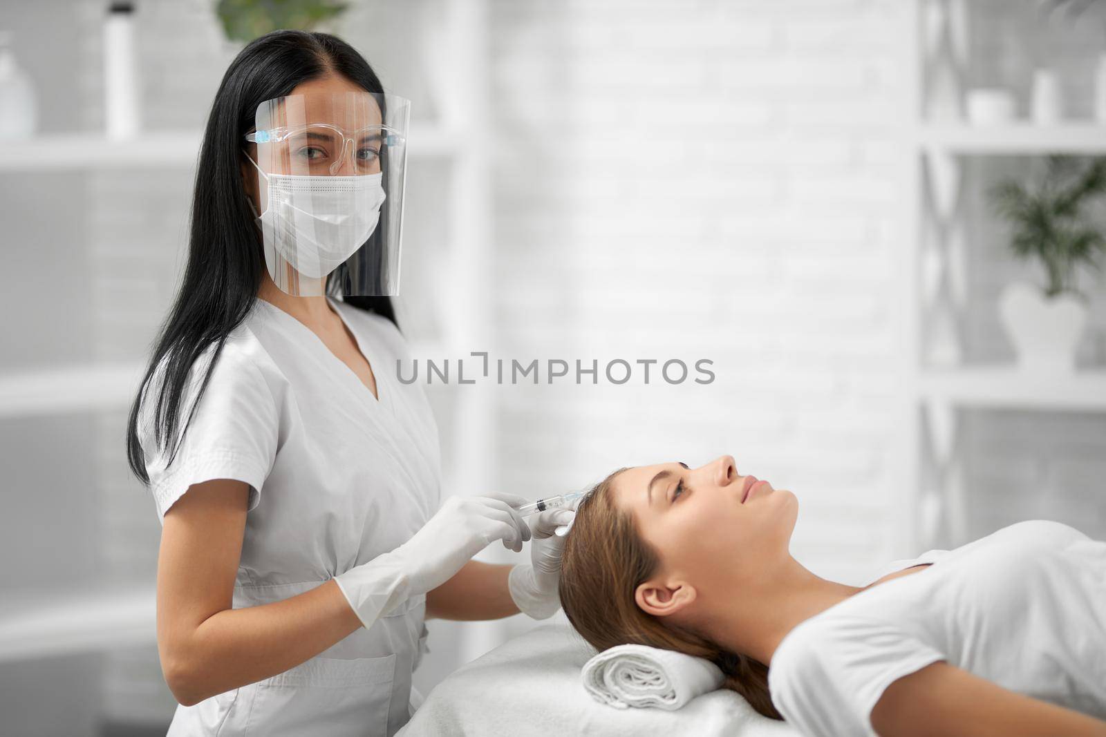 Side view of young brunette beautician in white rubber gloves doing injection for young cute woman in hair. Concept of improvements hair with special cosmetics.