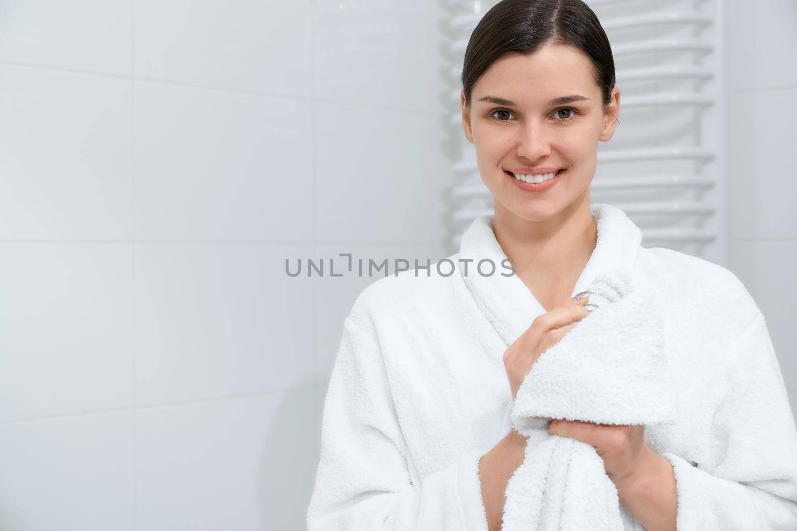 Front view portrait of smiling attractive young woman in white robe holding towel and wiping hands after shower. Concept of time for care body and hands.