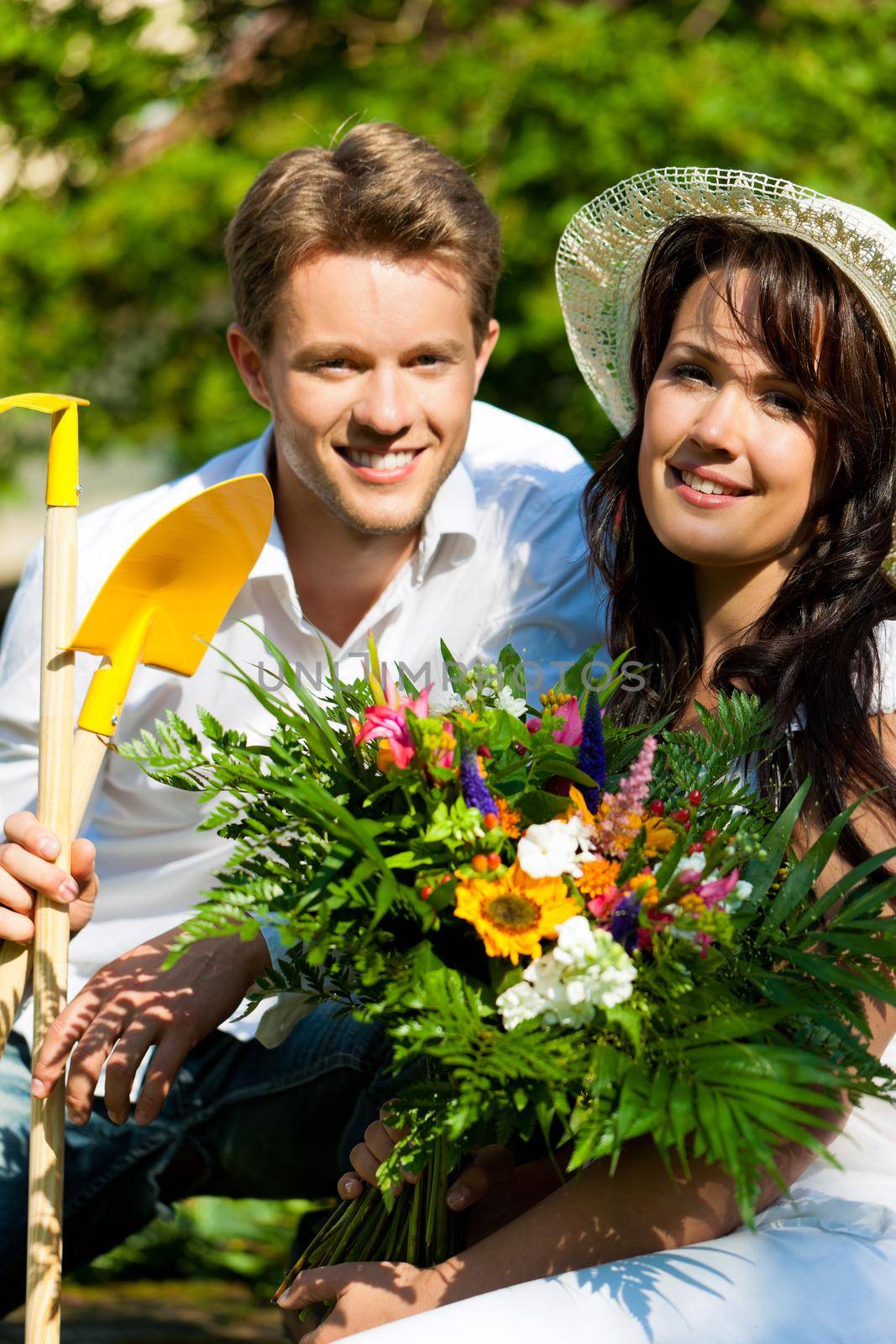 Happy couple gardening in summer with tools, she is wearing a hat
