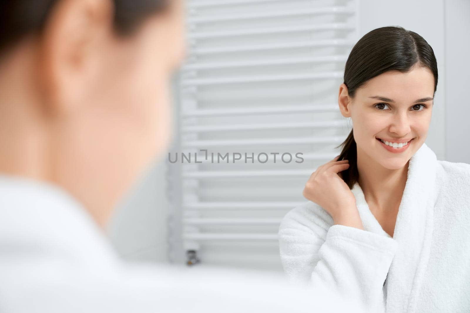 Close up portrait of smiling attractive young woman standing and looking at mirror in modern bathroom. Concept of time for different procedure for face and body with good mood. 