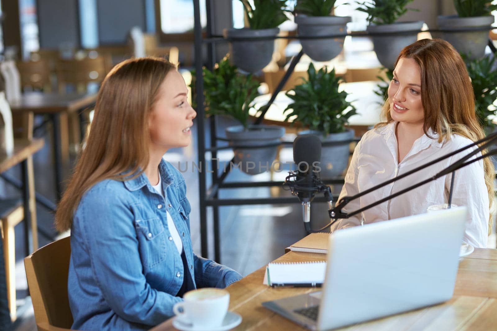 Side view of two beautiful young blogger women sitting in cafe and communicating with modern microphone and laptop. Concept of talking about different topics in cafe with tasty coffee. 