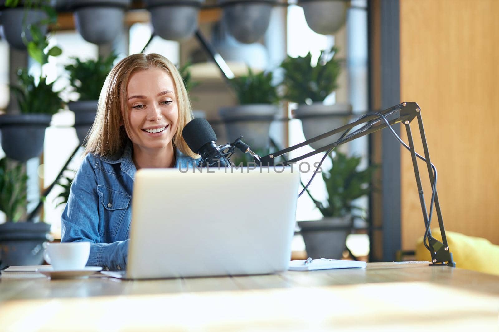 Front view of young cute woman with good mood communicating online in laptop with modern microphone in cafe. Concept of process working on radio with microphone and laptop. 