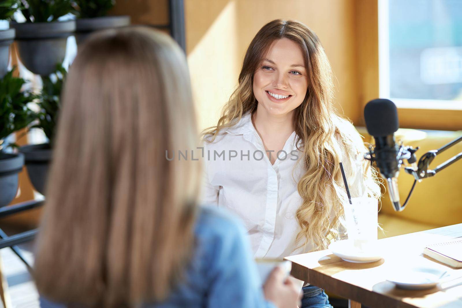 Side view of two attractive young women communicating in cafe with tasty coffee with good mood. Concept of process talking about different topics with modern microphone.