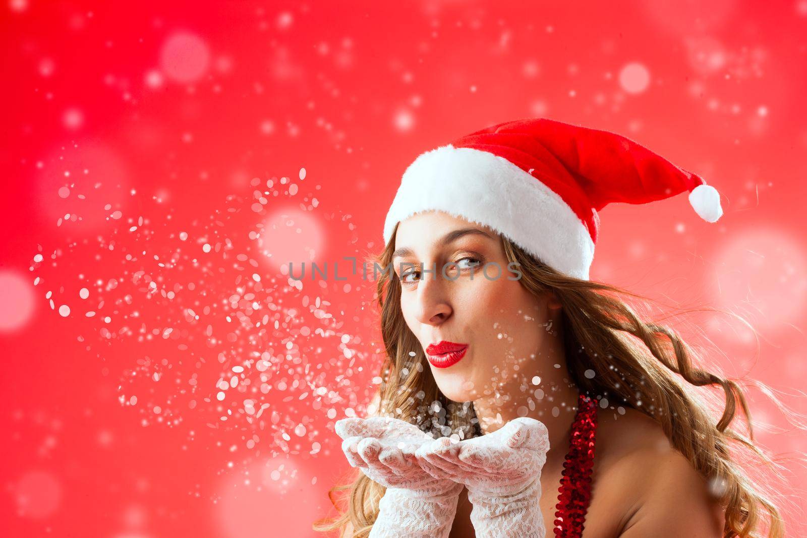 Attractive young woman in Santa Claus costume blowing snow flakes on isolated red background Xmas celebration