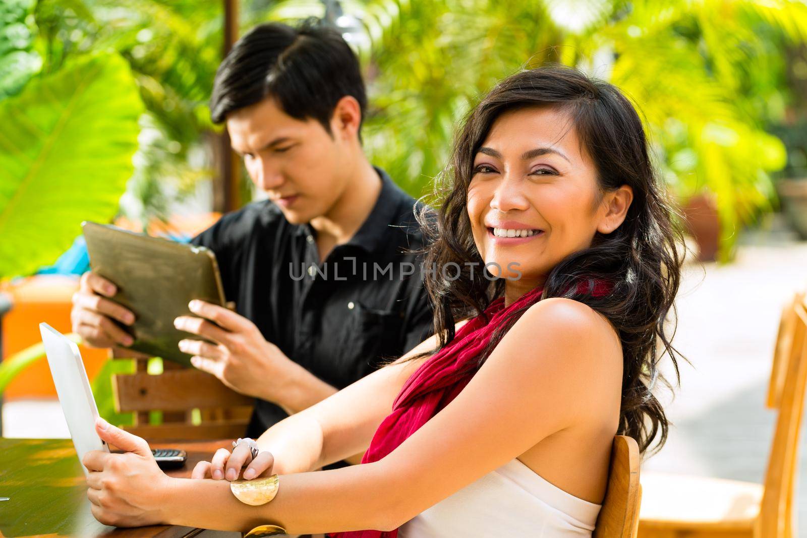 Asian woman and man are sitting in a bar or cafe outdoor and are surfing the internet with a tablet computer