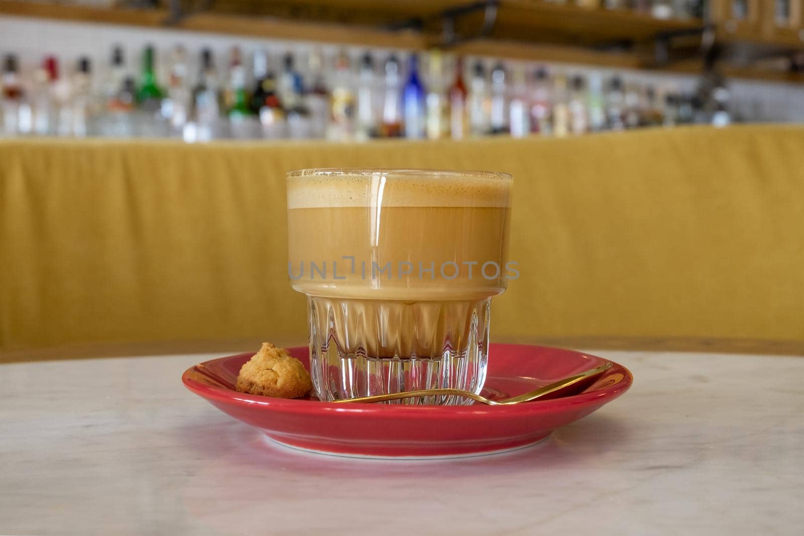 An elegant glass cup with flat white coffee served with a red saucer, golden spoon and small cookies. Coffee in the cafe. Selective focus, blurred background.