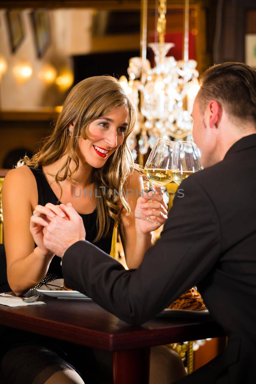 happy couple have a romantic date in a fine dining restaurant they drink wine and clinking glasses, cheers - a large chandelier is in Background