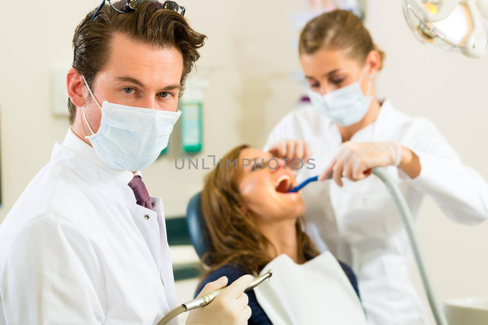 Dentists in his surgery looking at the viewer, in the background his assistant is giving a female patient a treatment