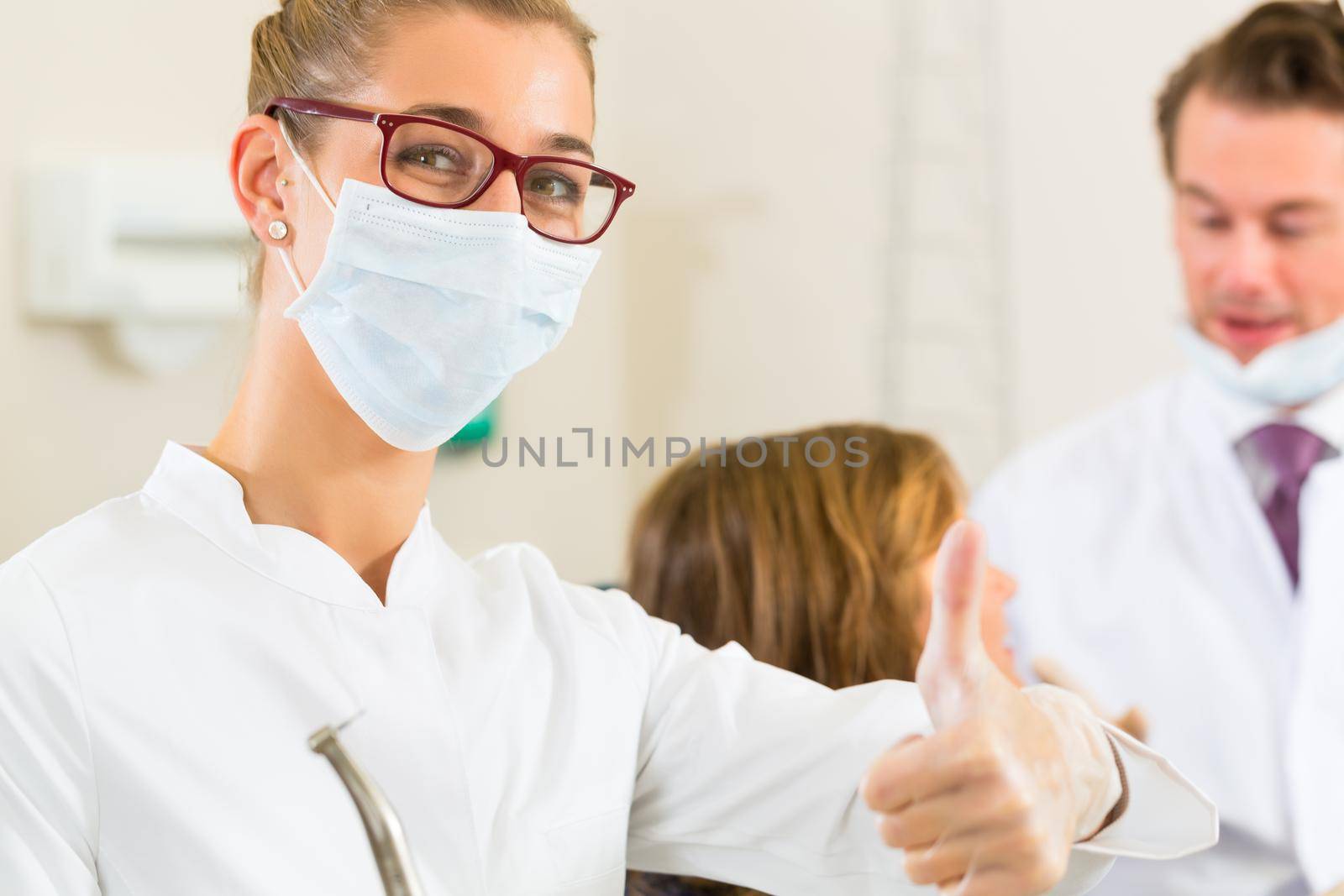 Dentists in his surgery holds a drill and looking at the viewer, in the background her colleague is giving a female patient a treatment