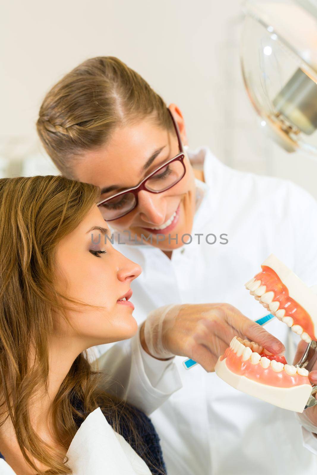 Dentist in his surgery holds a denture and explains a female patient with a toothbrush