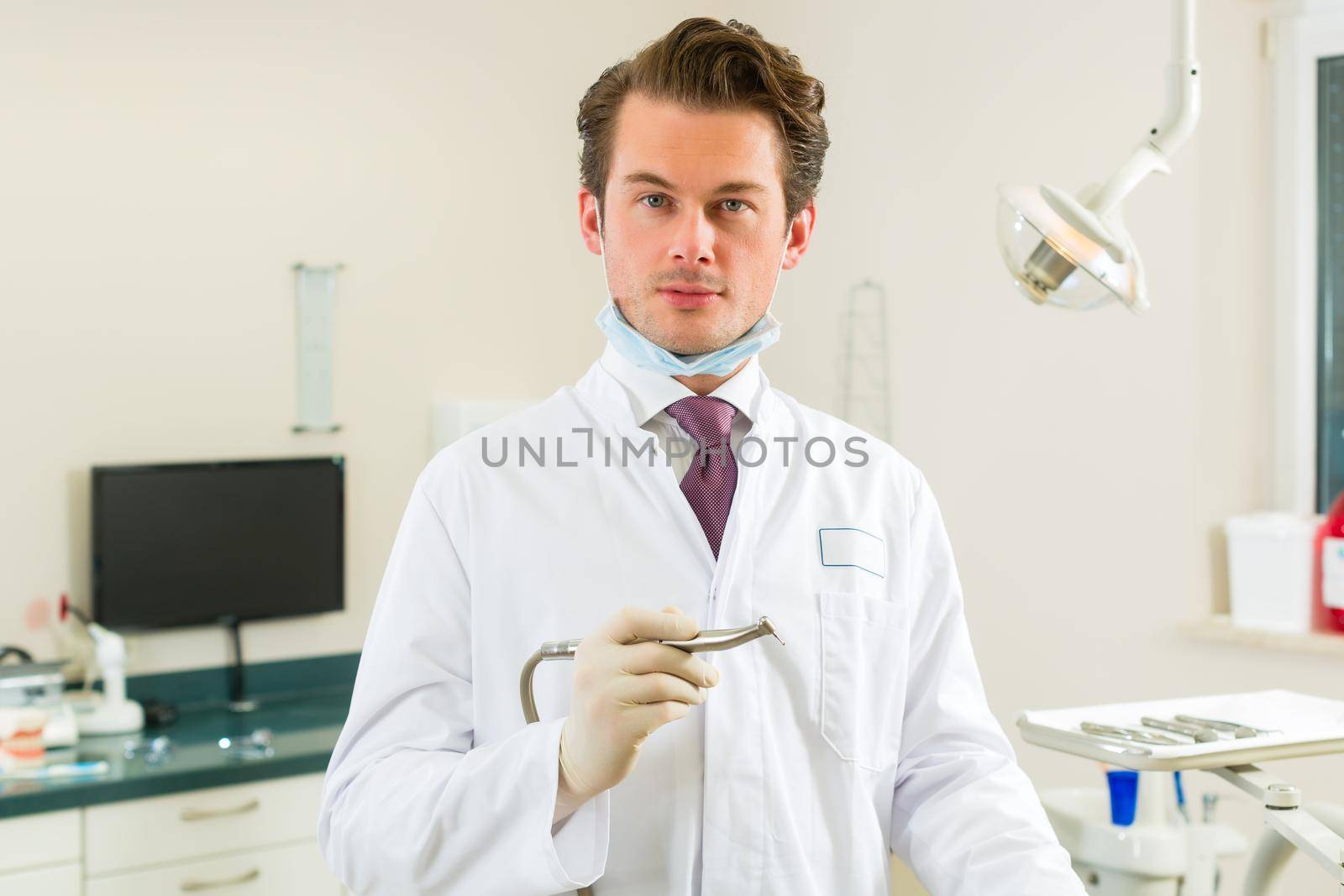 Dentists in his surgery holds a drill and looking at the viewer, in the background are tools for a dentist