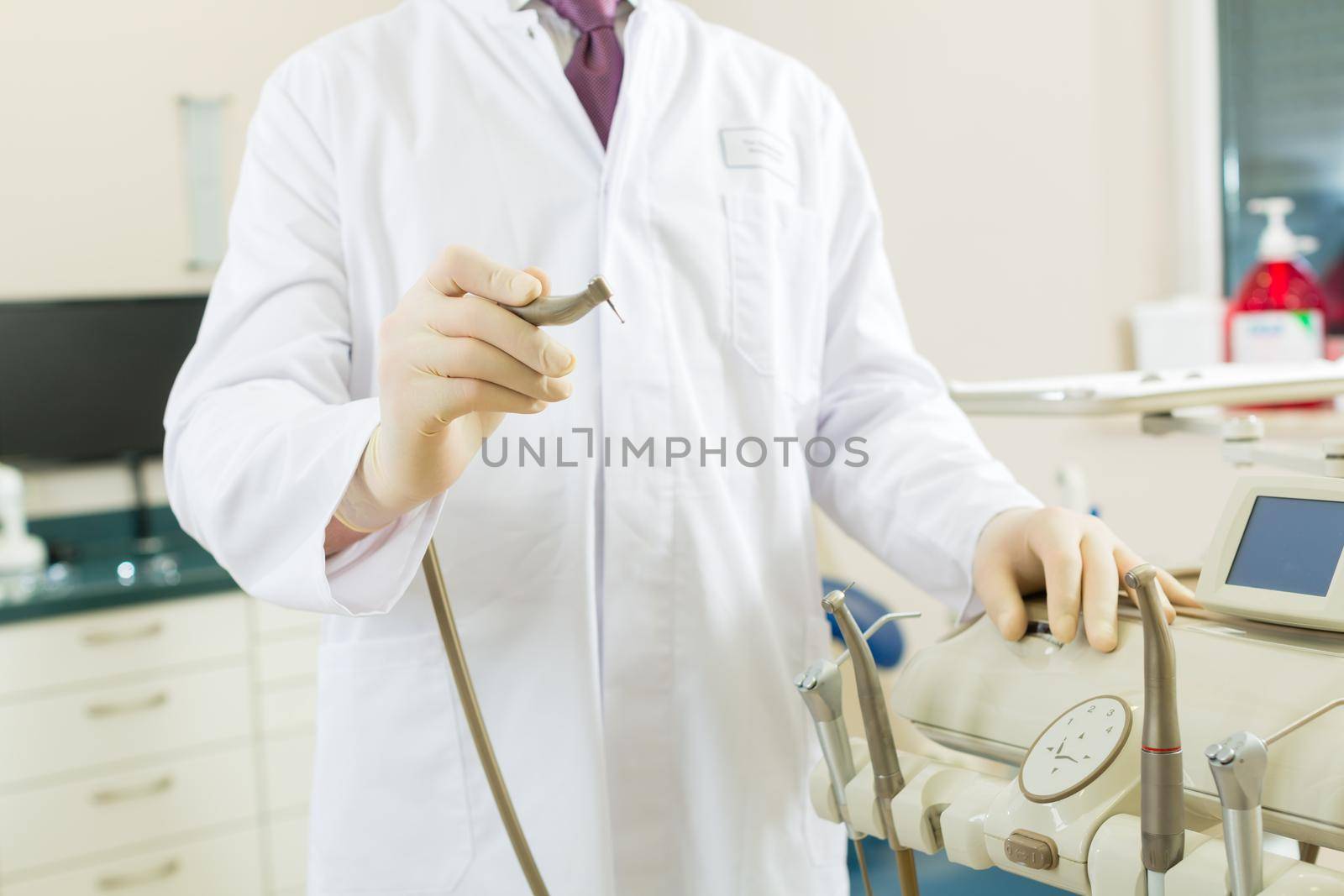 Dentists in his surgery holds a drill and looking at the viewer, in the background are tools for a dentist