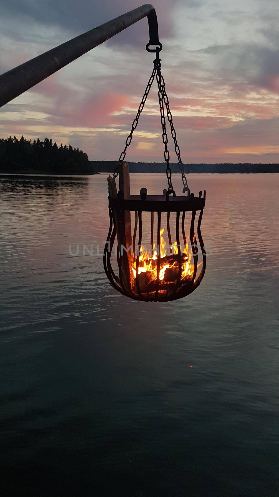 A bonfire burns in a metal basket hanging over the water at sunset