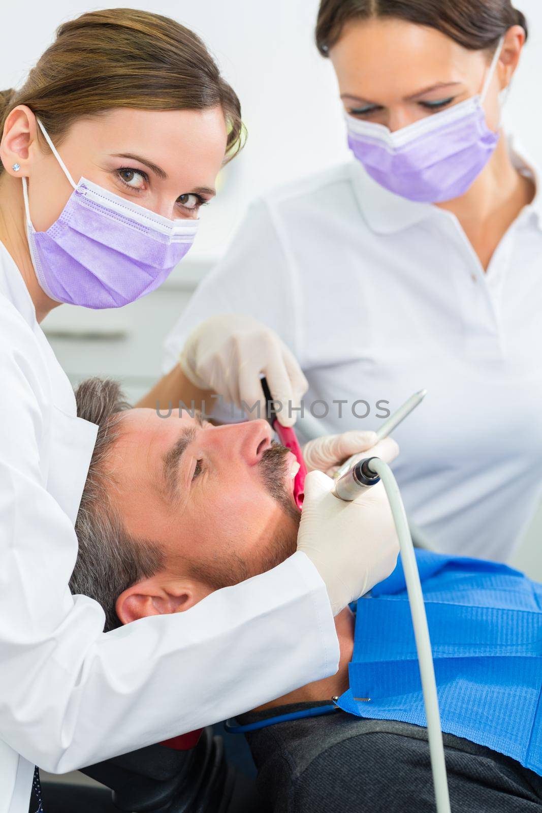 dentist in her practice or office treating male patient with assistant wearing masks and gloves