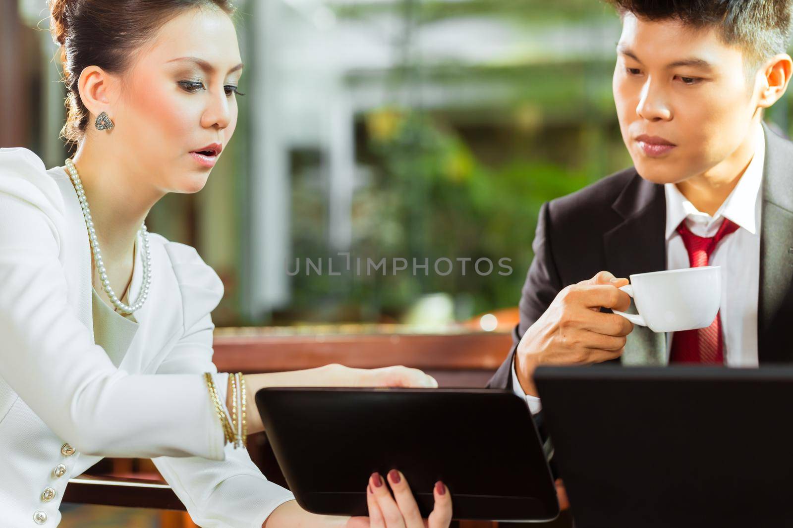 Two Asian Chinese office people or businessman and businesswoman having a business meeting in a hotel lobby discussing documents on a tablet computer while drinking coffee