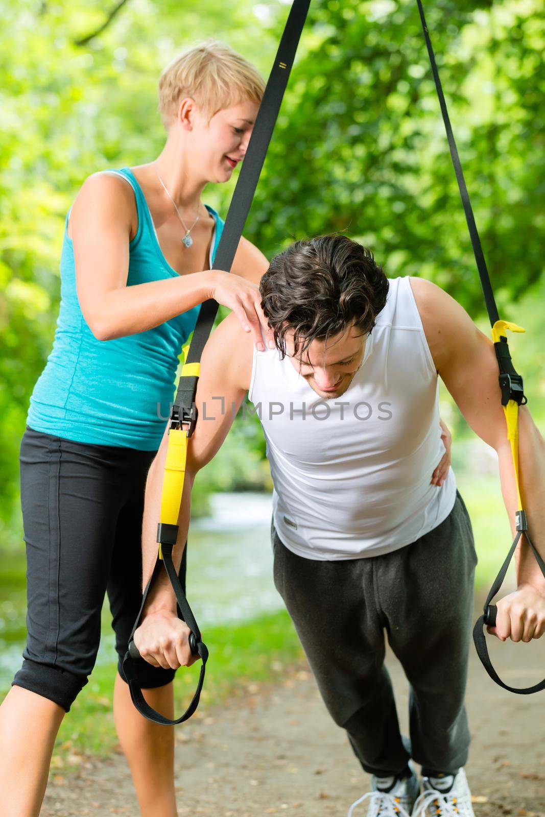 Young fitness man exercising with suspension trainer sling and personal sport trainer in City Park under summer trees