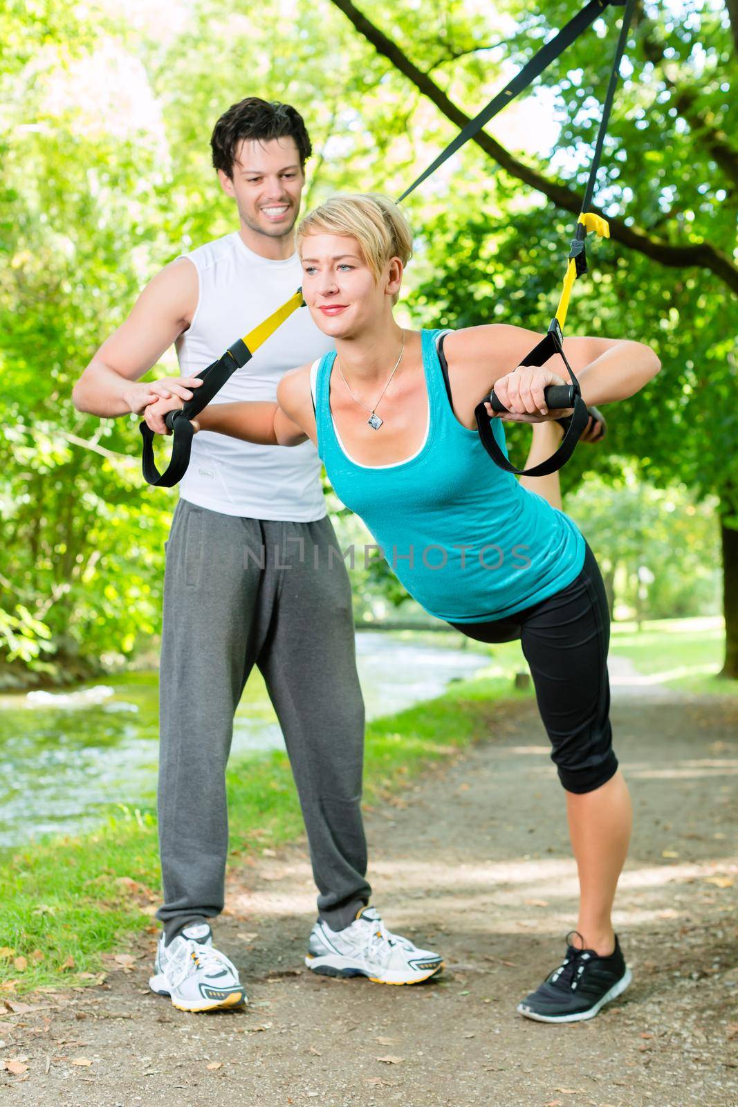 Fitness woman exercising with suspension trainer and personal sport trainer in City Park under summer trees