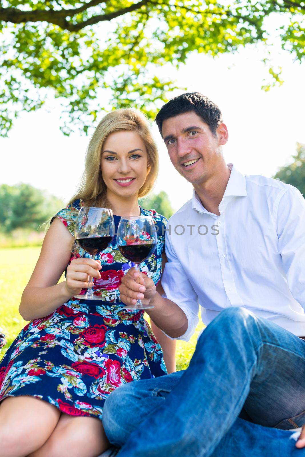 Couple having picnic wit red wine on meadow sitting on blanket with basket