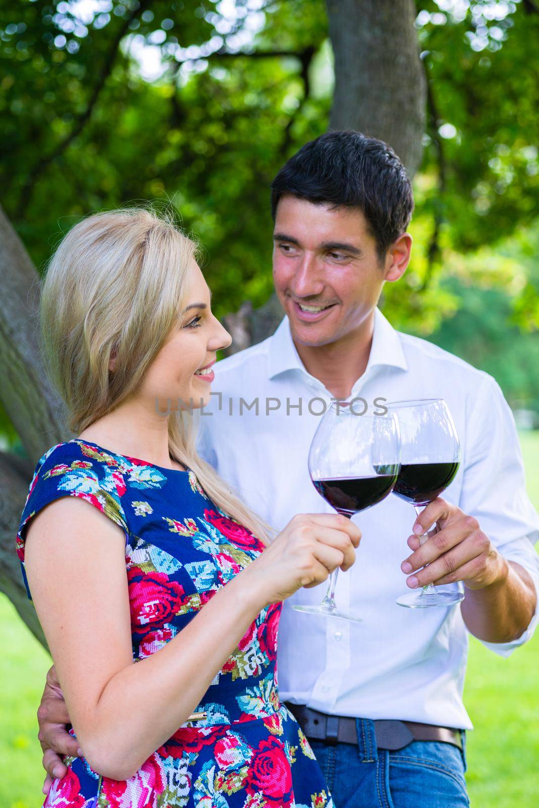 Couple having picnic wit red wine on meadow sitting on blanket with basket