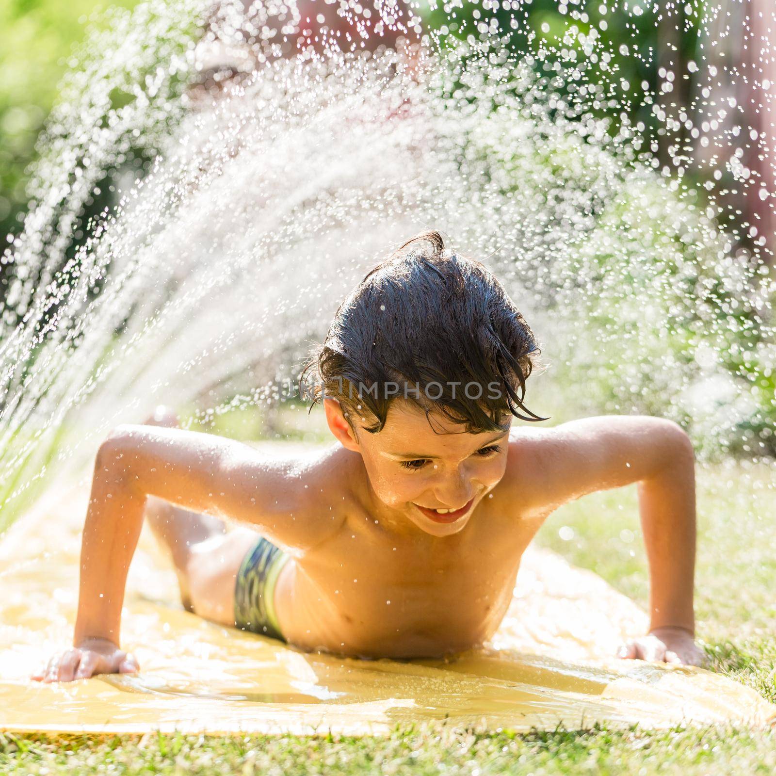 Boy cooling down with garden hose, family in the background on a hot summer day