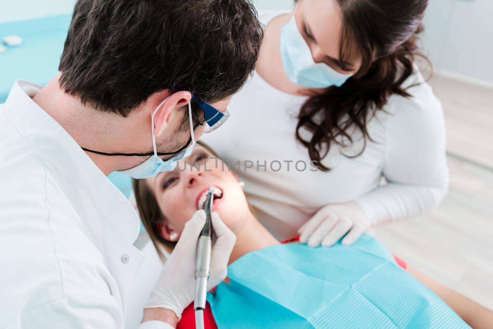 Dentist and assistant treating woman in clinic using drill on her teeth