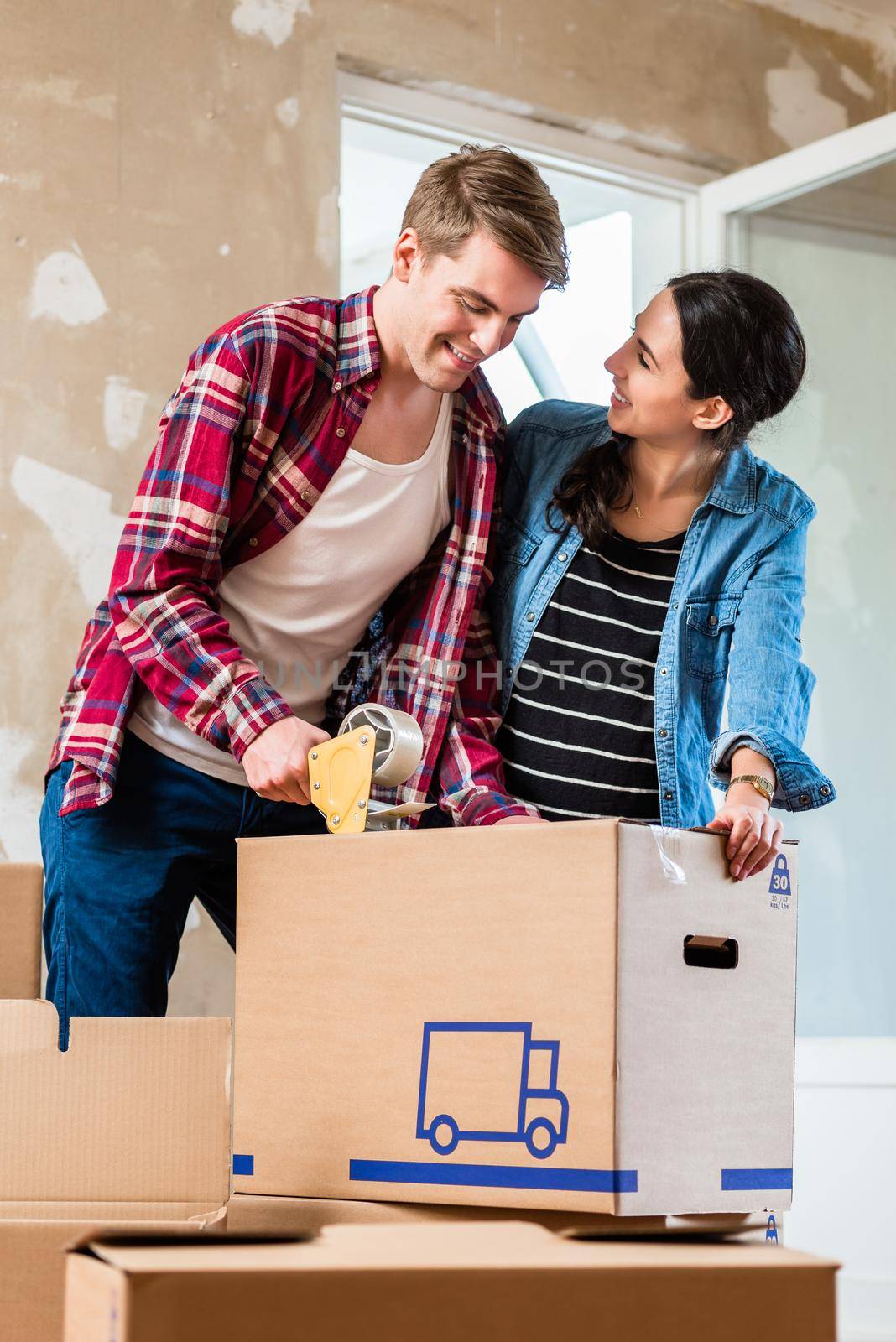 Young man and his girlfriend sealing a brown cardboard box with adhesive tape while renovating together their new home