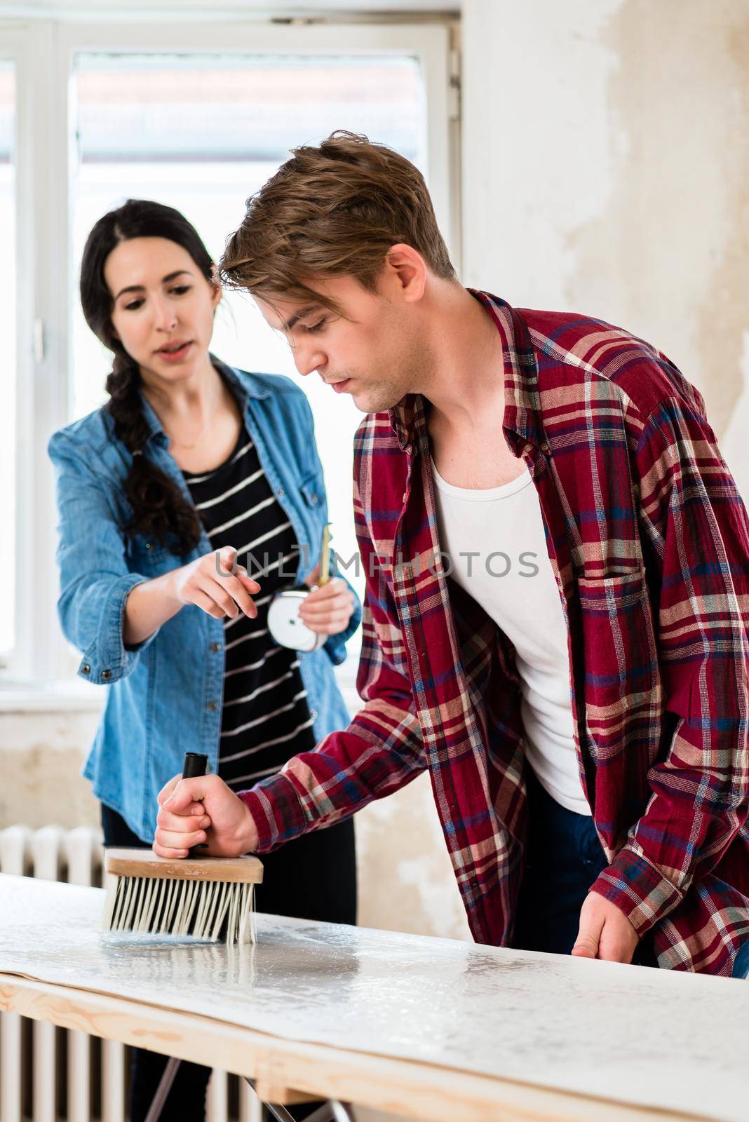 Funny young couple in love sharing tools for wallpapering while working together to the improvement of their new house