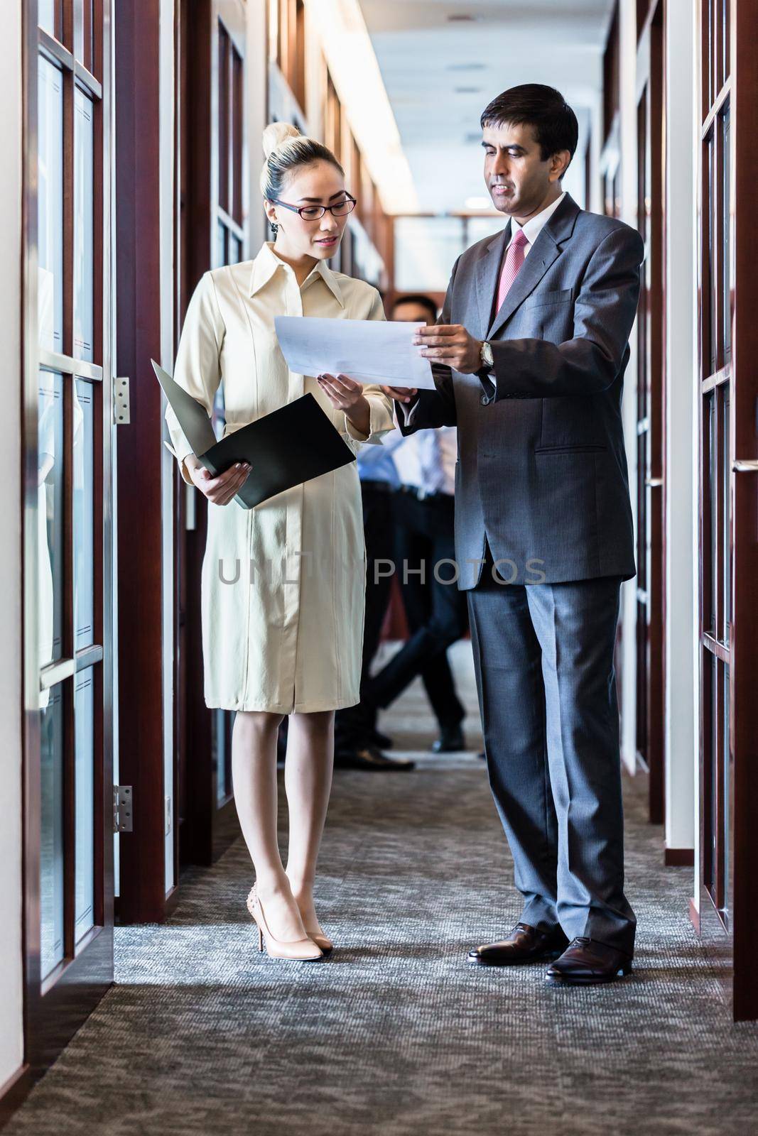 Indian business executive man and Indonesian secretary standing in office hallway discussing papers