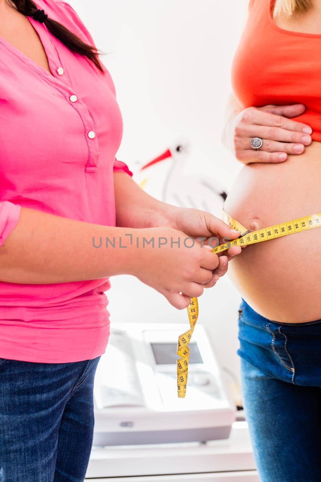 Standing pregnant women and midwife measuring circumference of baby bump with ribbon