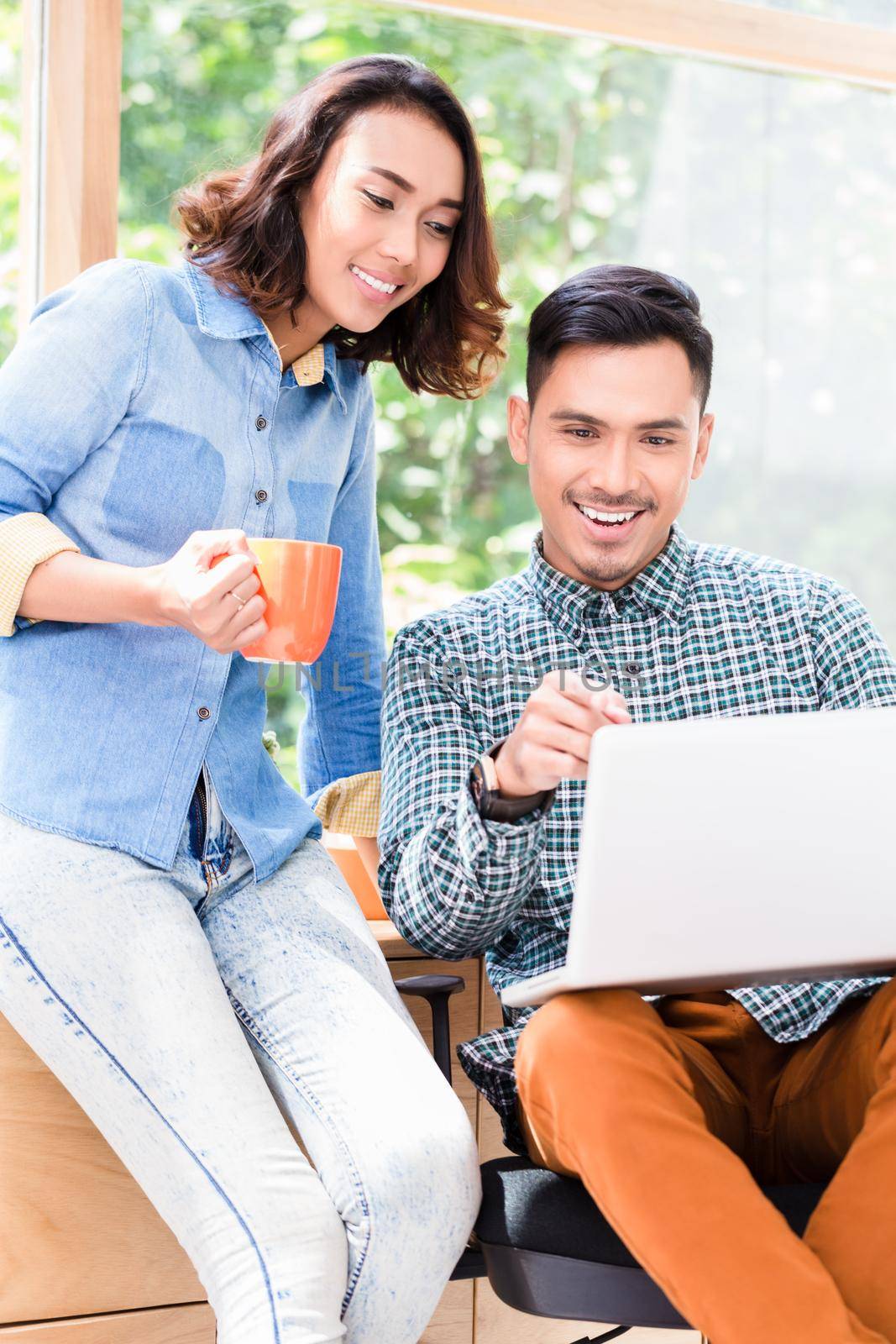 Young happy Asian employee looking at his colleague while sitting at desk in a relaxed working environment