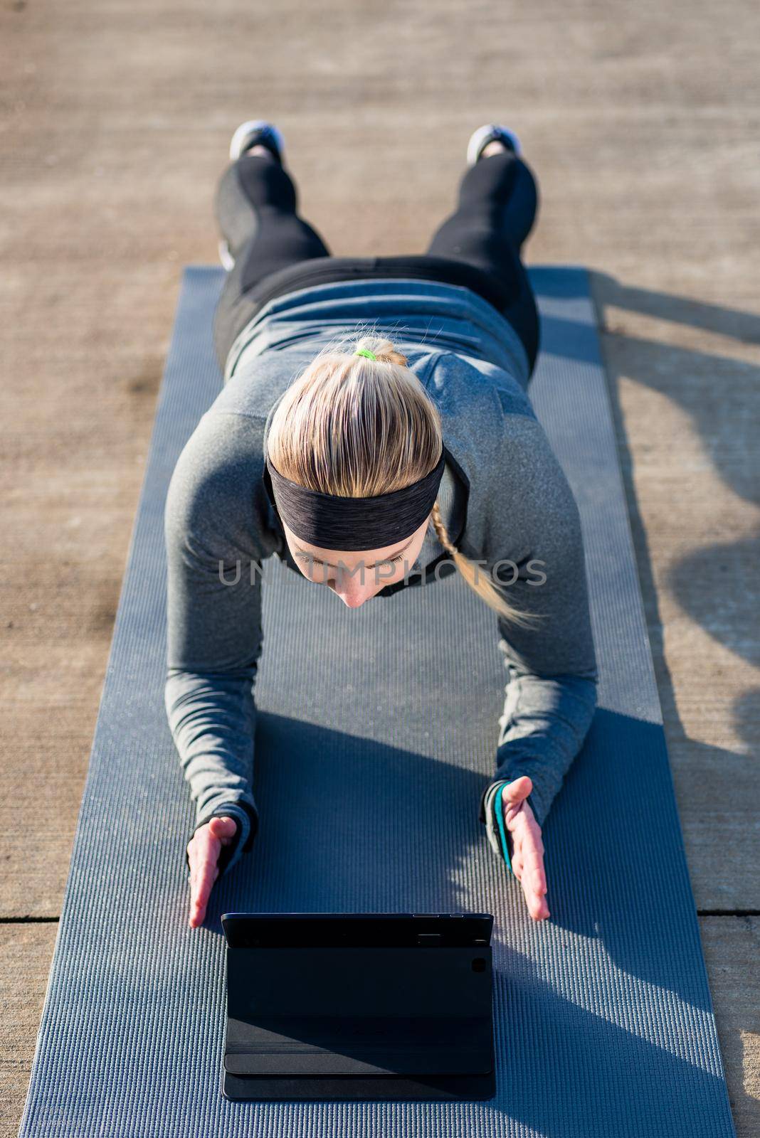 High-angle view of a fit young woman, watching a motivational video on tablet PC while exercising the forearm plank position on a mat outdoors in the park