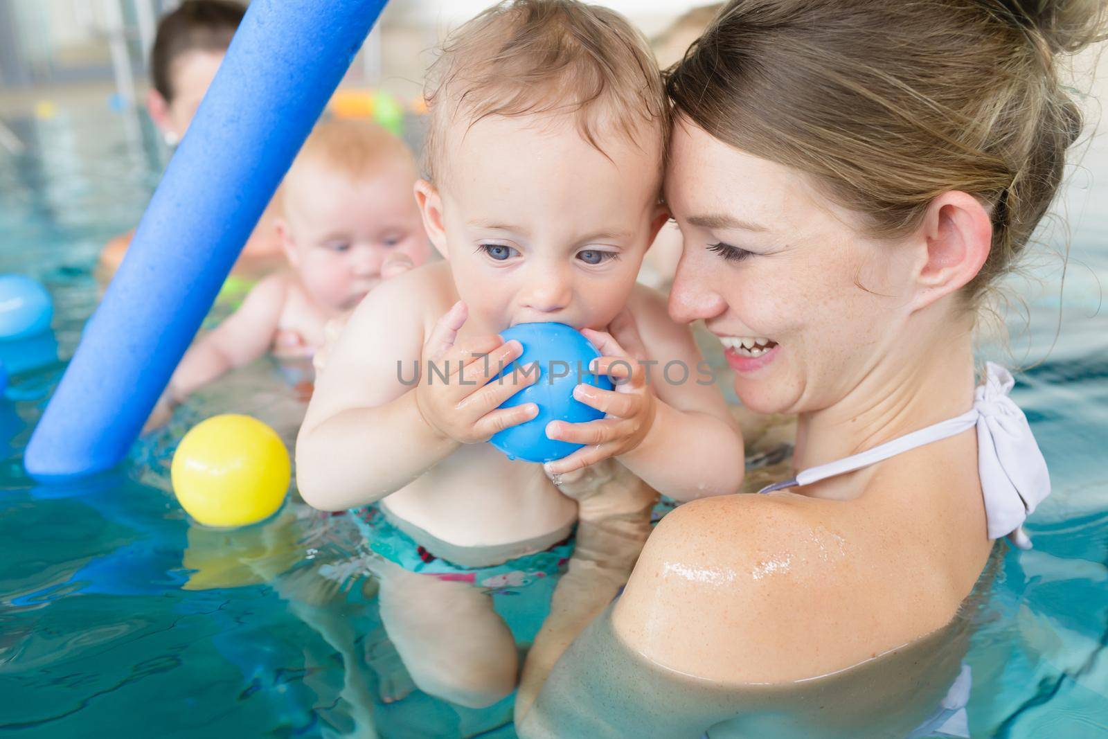 Mothers and their kids having fun at baby swimming lesson between lots of water balls