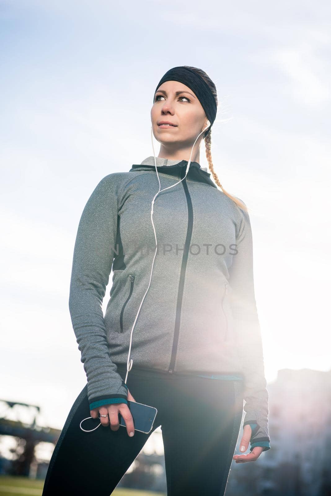 Beautiful thoughtful young woman listening to music on her mobile phone while wearing trendy sport outfits in a sunny day in the park