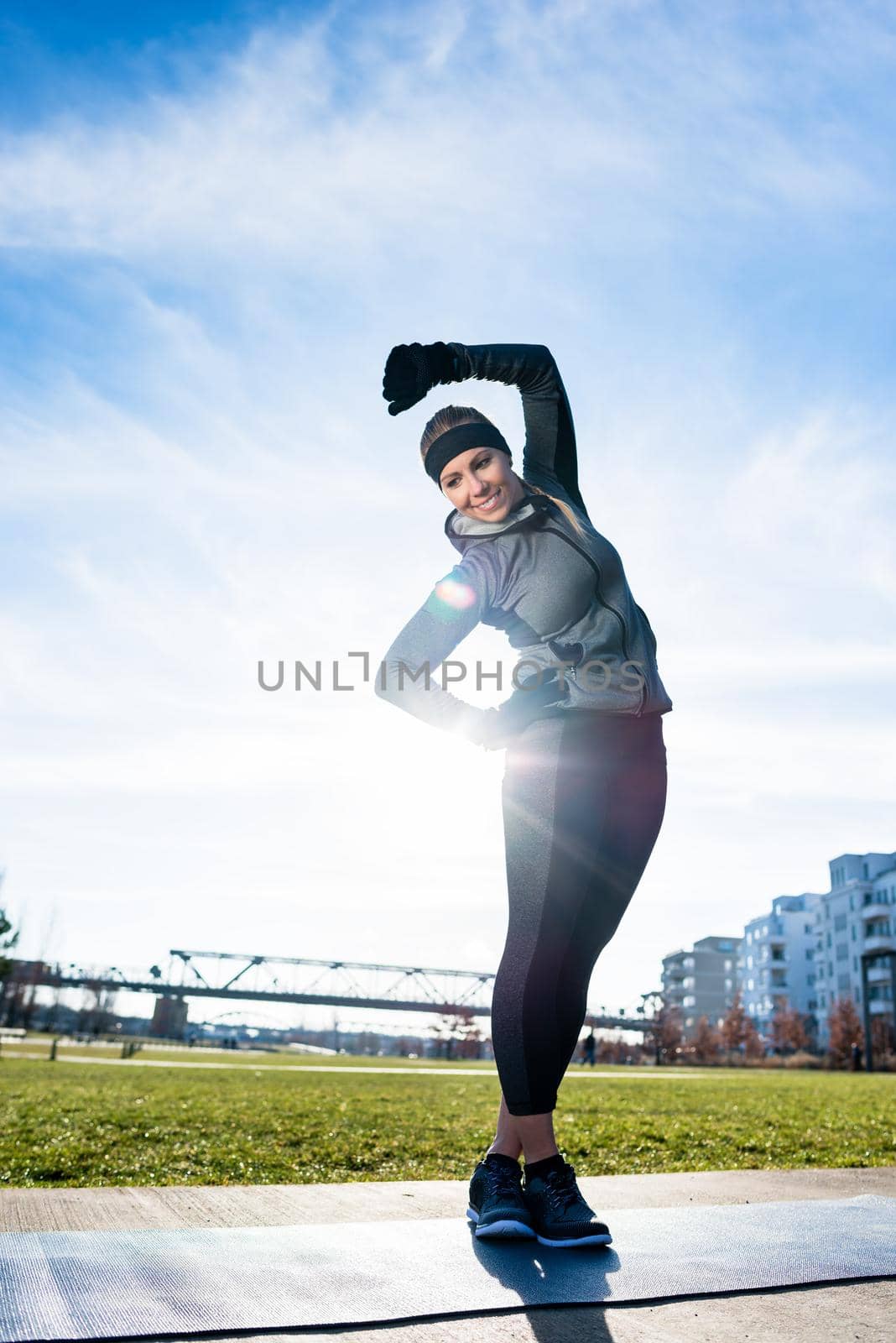 Athletic young woman practicing stretching exercise while standing with crossed legs outdoors in a sunny day in the park