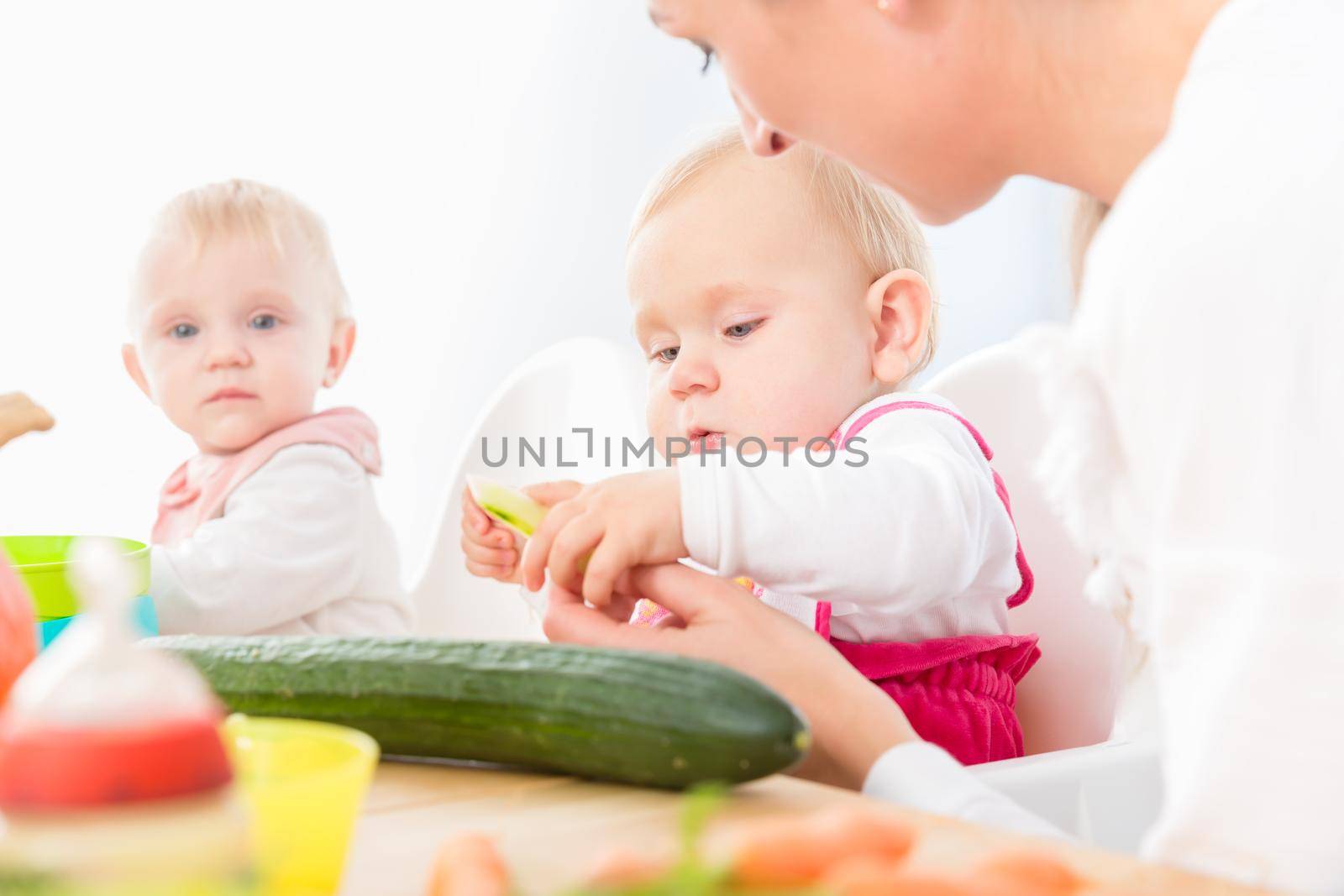 Portrait of a cute baby girl with blue eyes eating healthy solid food, while sitting in a high chair next to another baby in a modern daycare center for babies