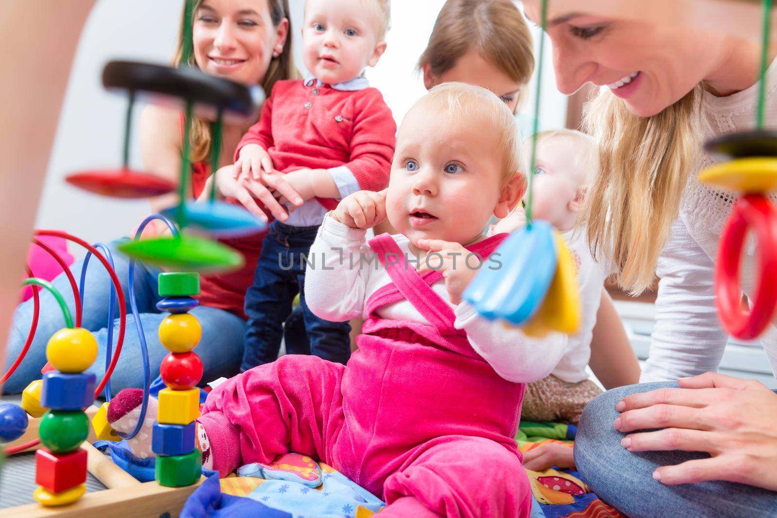 Portrait of a cute baby girl looking up with her beautiful blue eyes, while sitting down on the floor with other two babies and their mothers at home