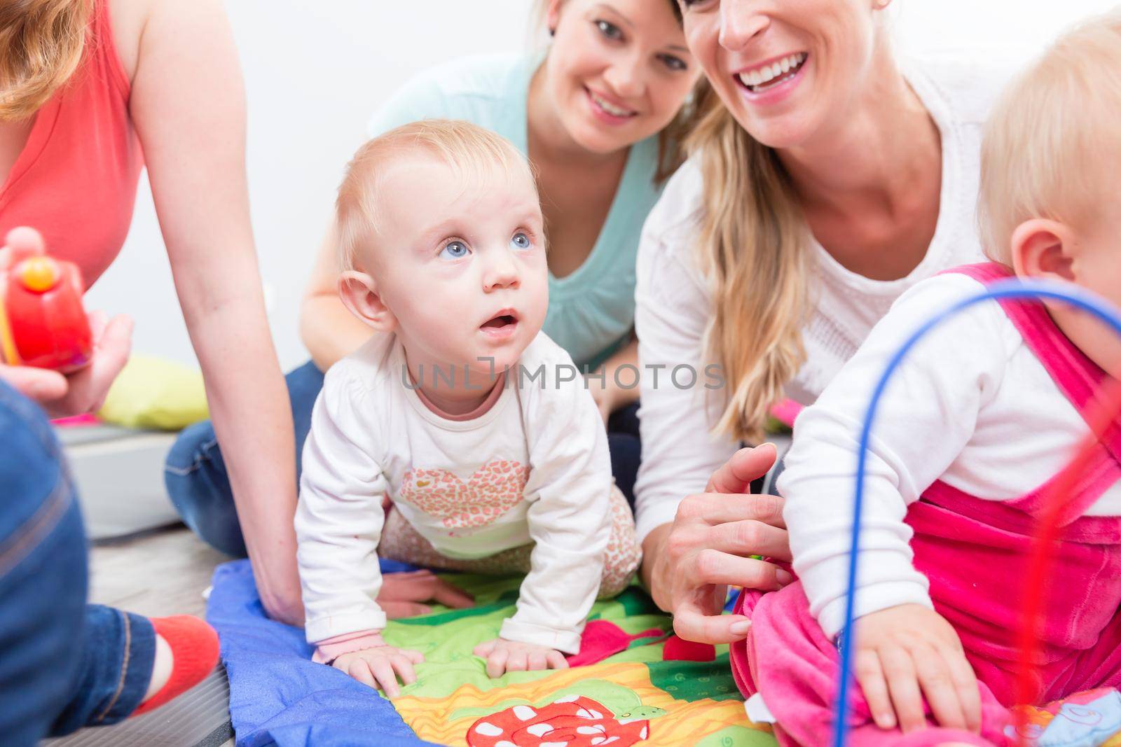 Group of happy young mothers watching their cute and healthy babies while playing with multicolored toys in a modern daycare center