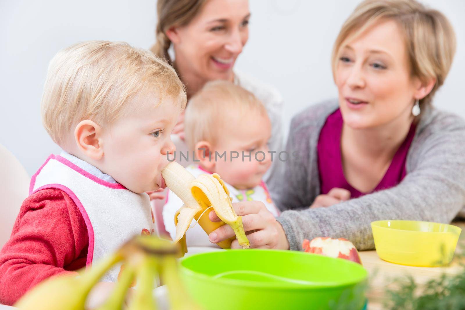 Portrait of a cute and healthy baby girl with blue eyes looking at her mother, while eating nutritious fruit puree next to another baby at home