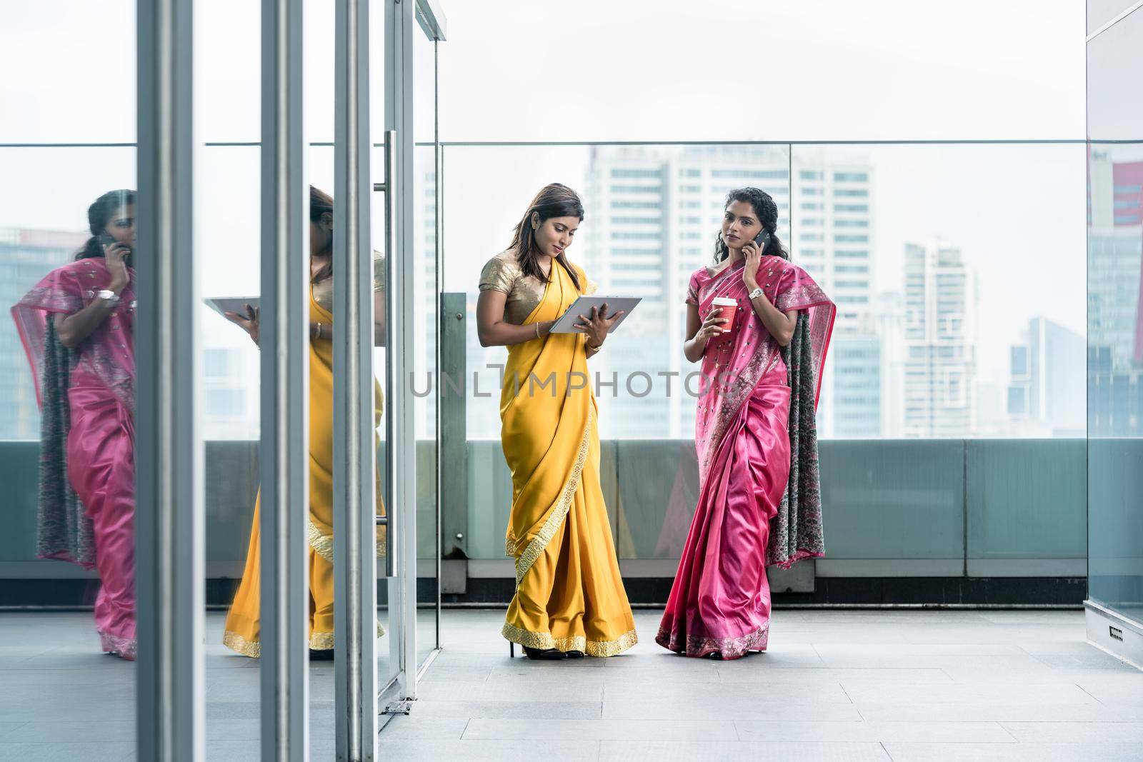 Two young Indian women using modern technology for communication during the break at work in a business building