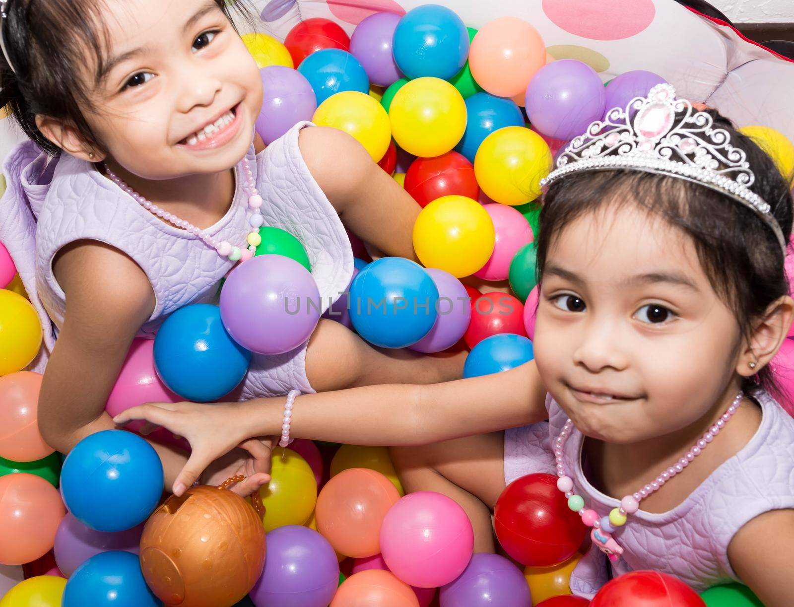 Female asian identical twins sitting on chair with white background. Wearing purple dress and accessories crown. Playing colorful plastic toy balls