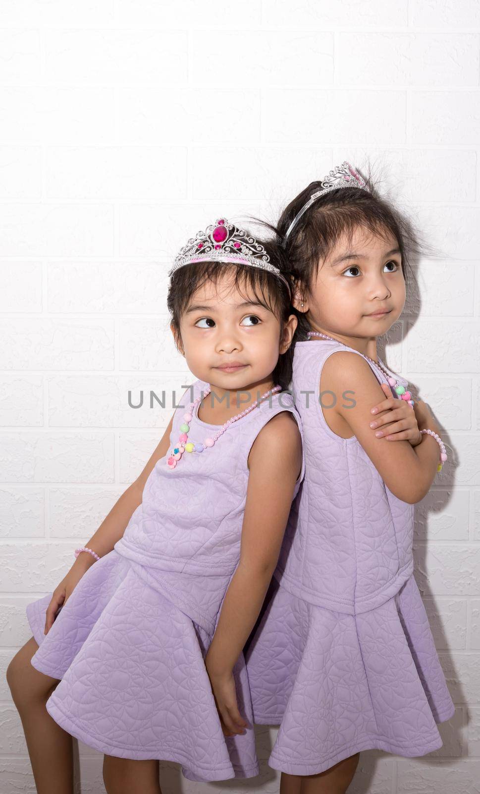 Female asian identical twins sitting on chair with white background. Wearing purple dress and accessories. Standing back to back with each other and having fun