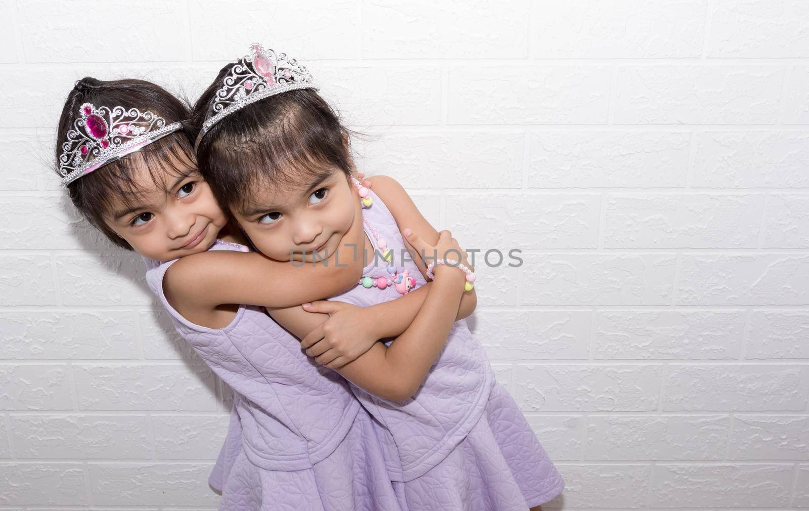 Female asian identical twins sitting on chair with white background. Wearing purple dress and accessories. Standing hugging each other by billroque