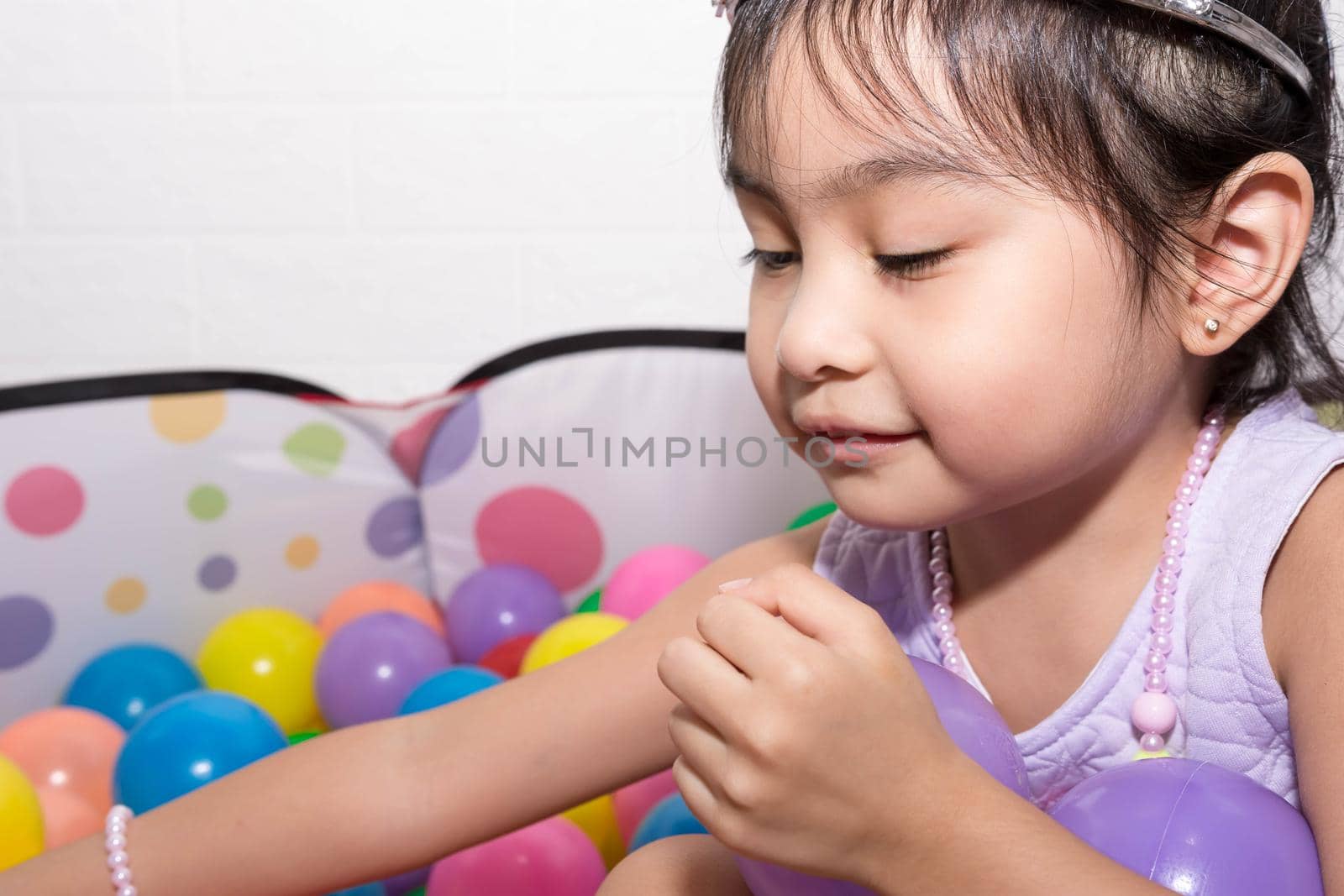 Female asian child girl  while sitting and playing with colorful plastic balls while wearing accessories like crown and necklace